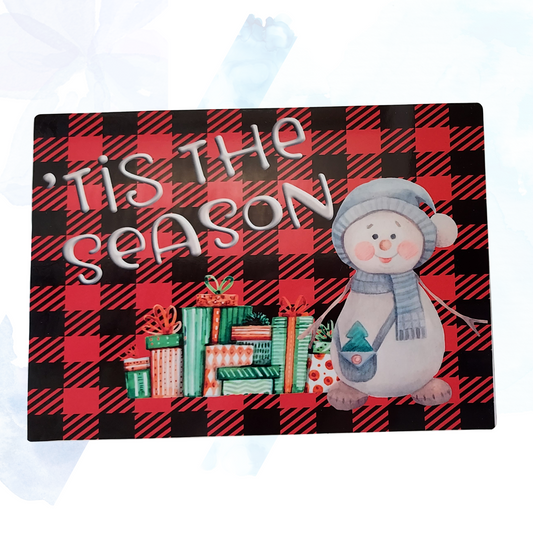 A 5" x 7" shelf sitter home decor sign with a festive design. The sign features a cheerful snowman and wrapped presents set against a red plaid background. The text "'Tis the Season" is displayed in elegant white script, adding a charming holiday touch to any room. This compact and festive sign is perfect for adding seasonal cheer to shelves, mantels, or tabletops.